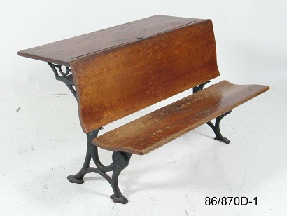 Dual seat wooden school desk with cast iron legs and pen rest and centre ink well on desktop.